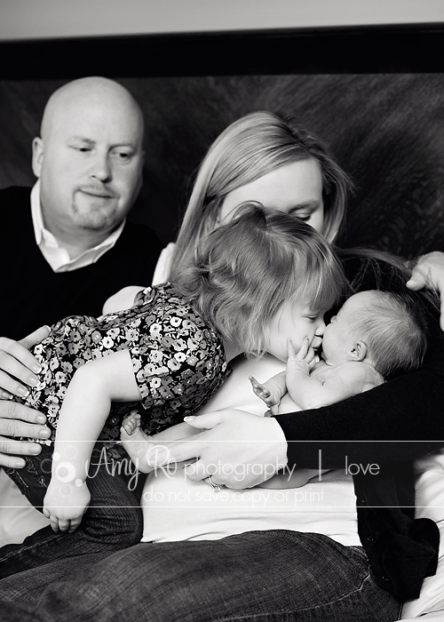 Family image with big sister kissing newborn on bed