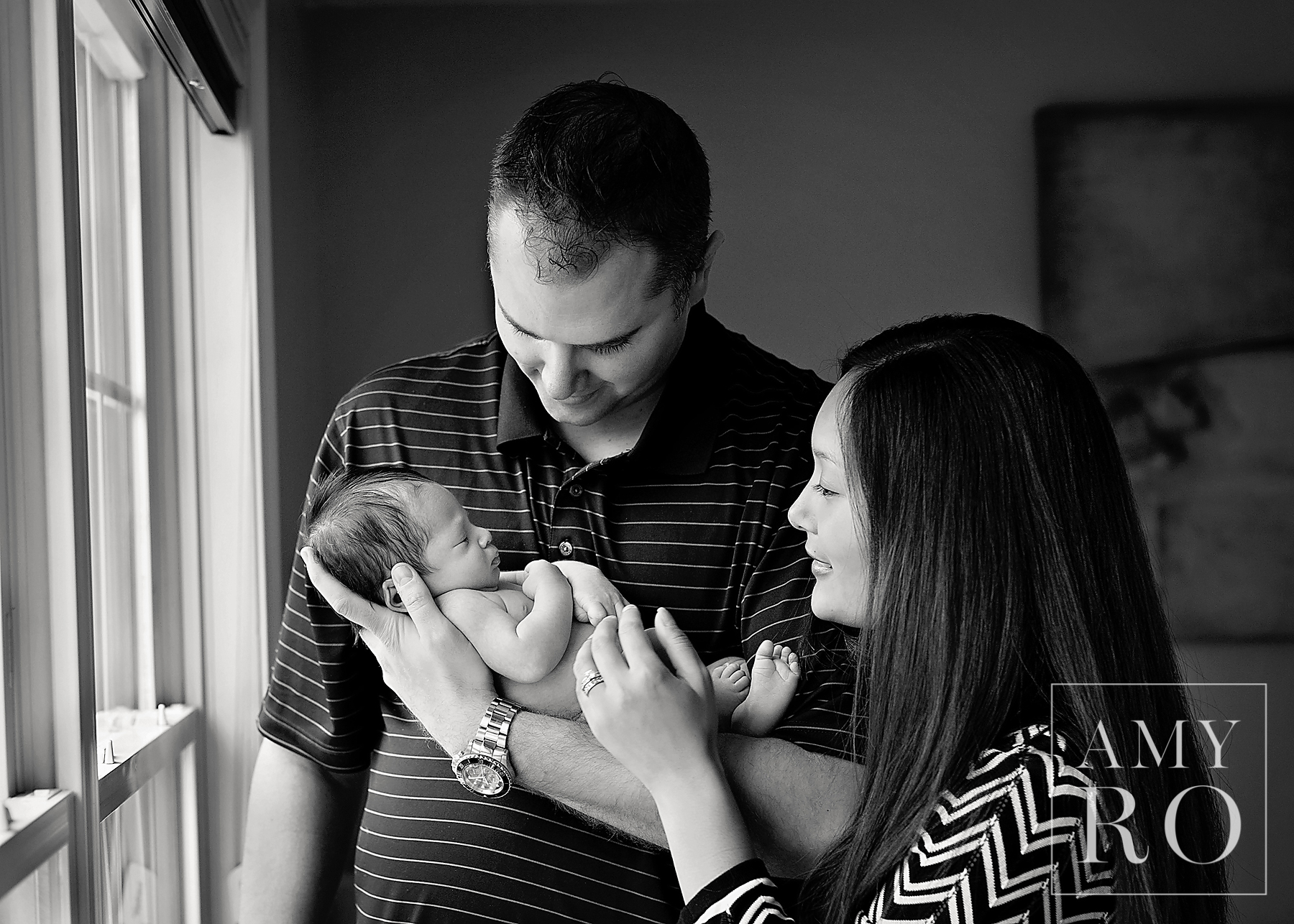Black and white image of mom, dad and newborn baby boy in the bedroom by a window taken during a Massachusetts newborn photography session