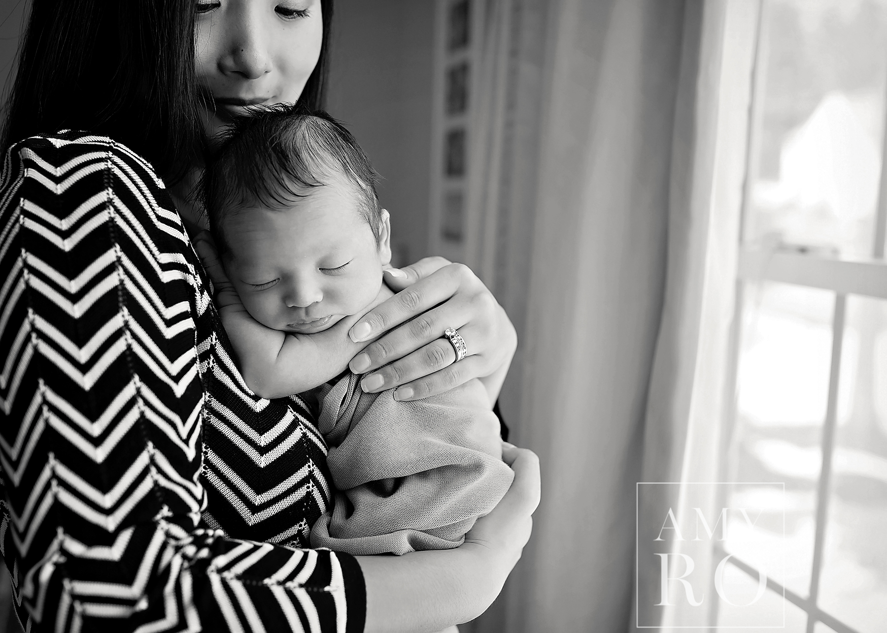 Black and white image of mom holding newborn next to the nursery window, sleeping newborn boy