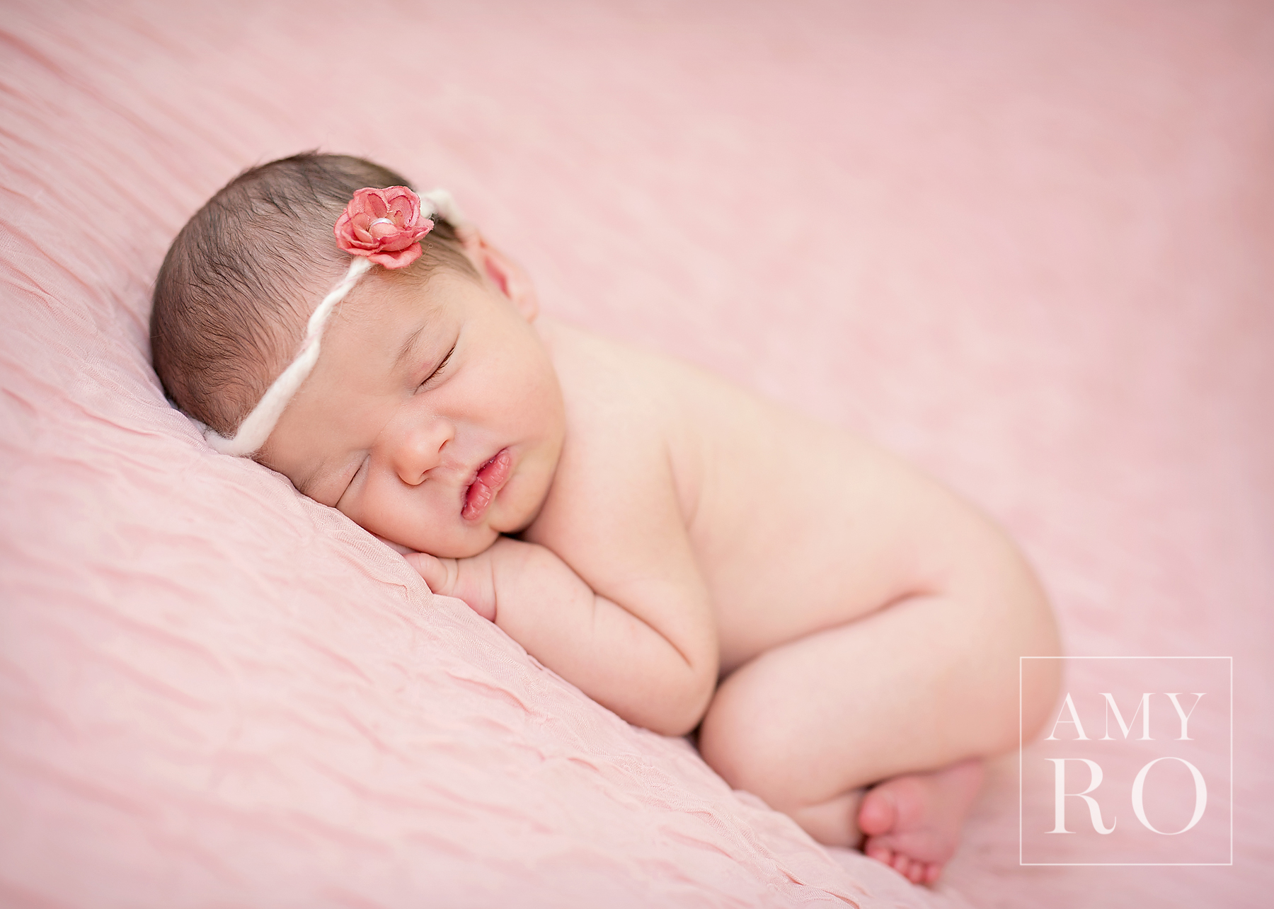 Beautiful image of newborn girl on pink backdrop with flower head band curled and sleeping during a newborn photography lifestyle session in Rhode Island