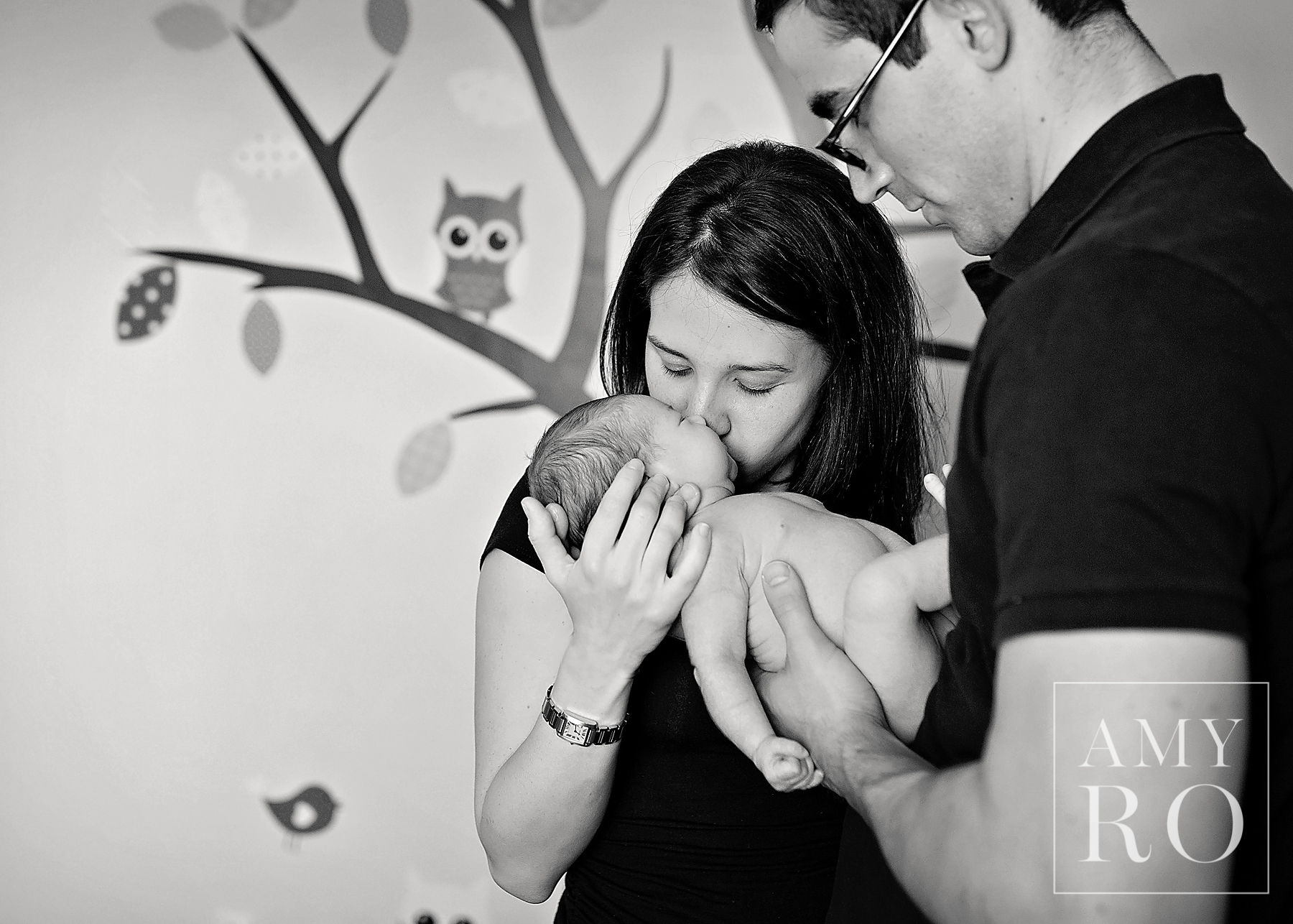 Black and white image of mom and dad snuggling baby in the nursery, with mom kissing baby taken during newborn session in Rhode Island