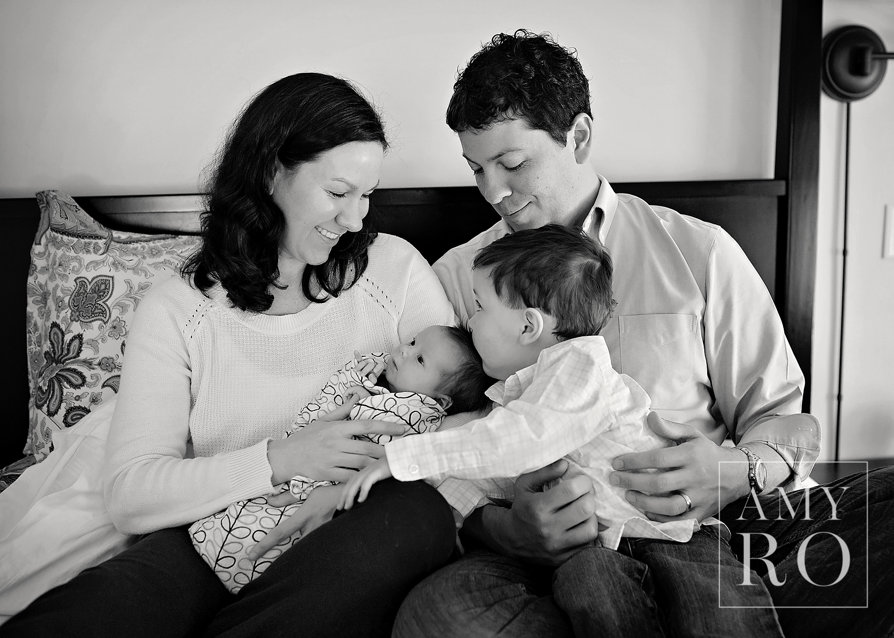 Black and white image of family snuggled on the bed with big brother kissing newborn sister taken during  lifestyle newborn session in Massachusetts