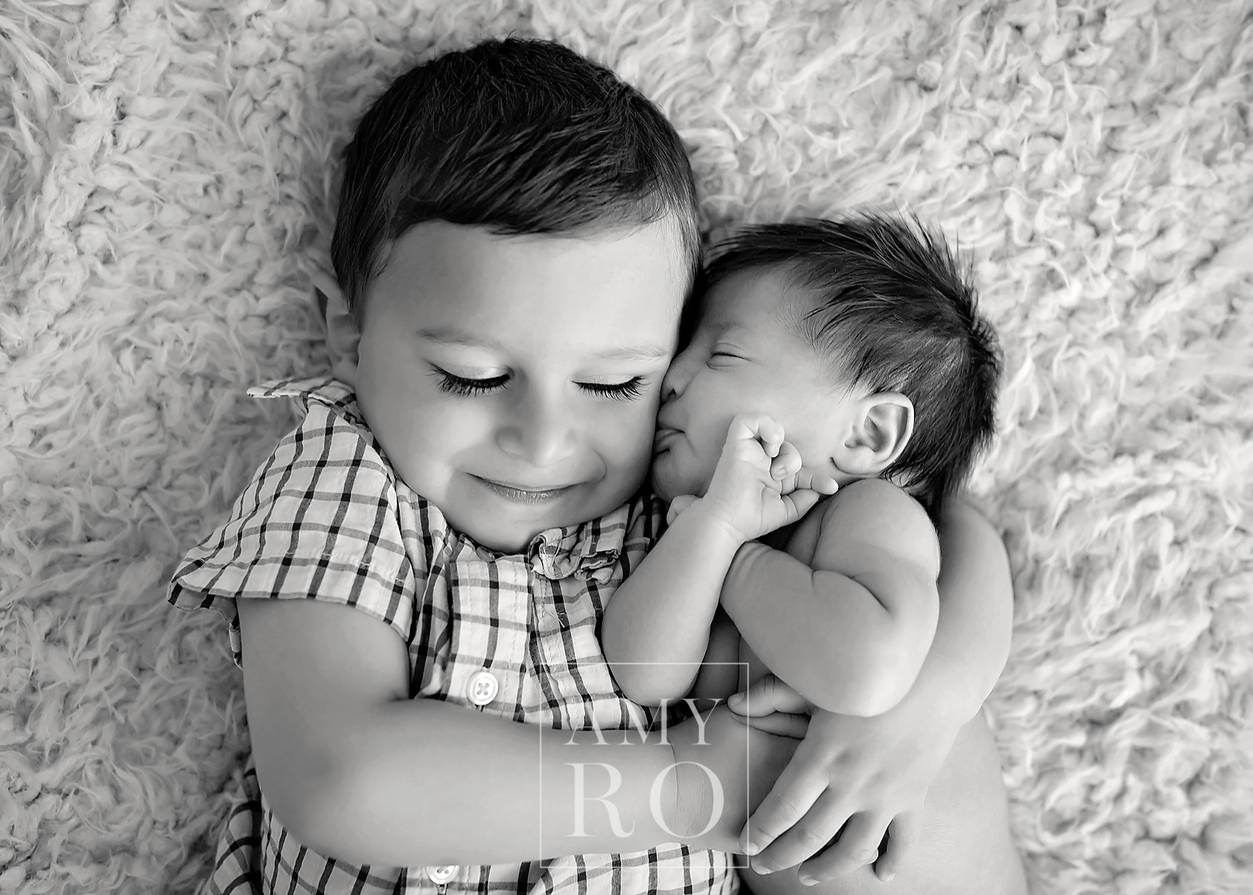 Black and white image of brother cuddling with newborn baby sister with eyes closed during a newborn session in Rhode Island, sibling newborn kisses