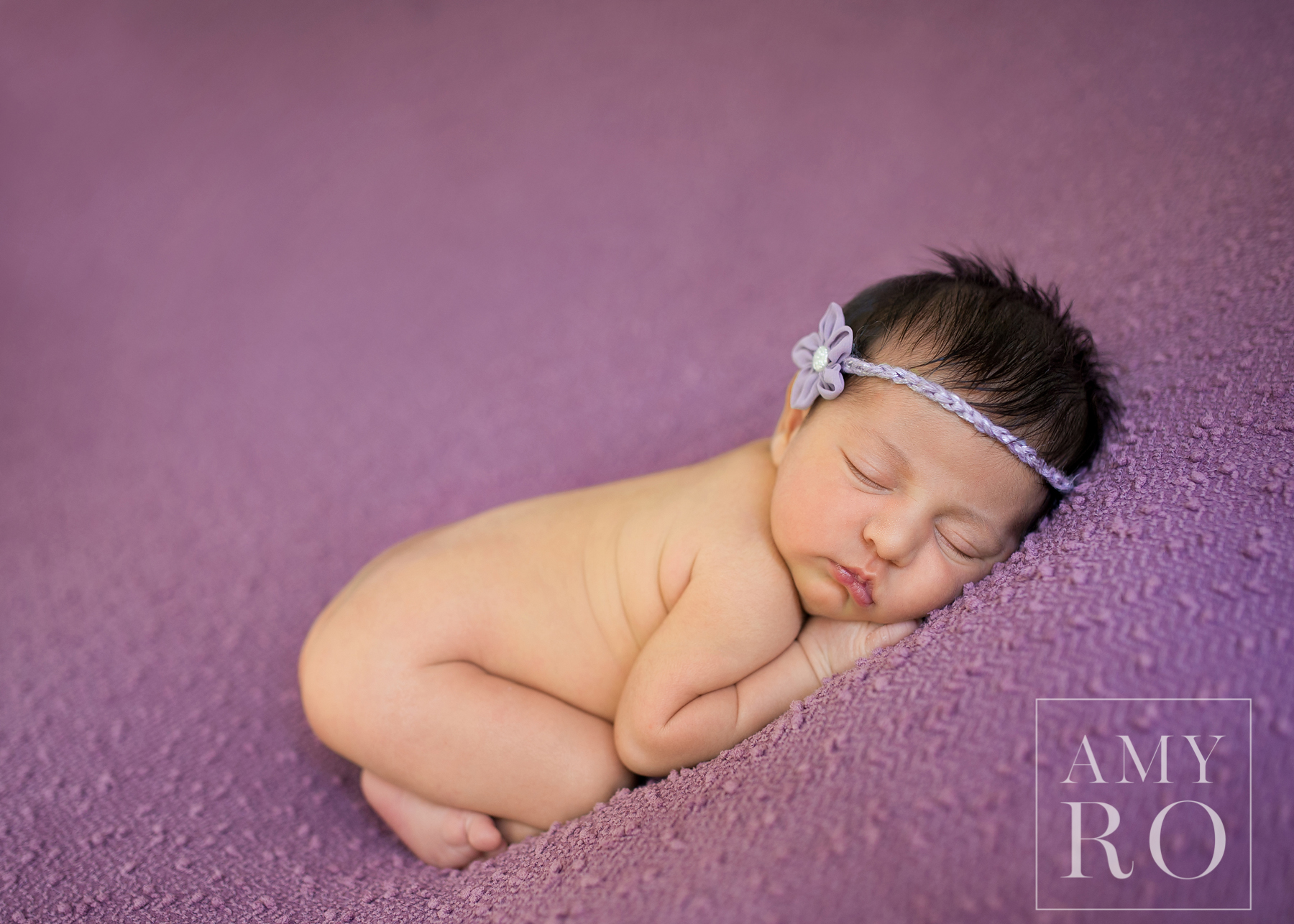 Newborn girl curled up sleeping on a purple blanket, brown hair, purple headband
