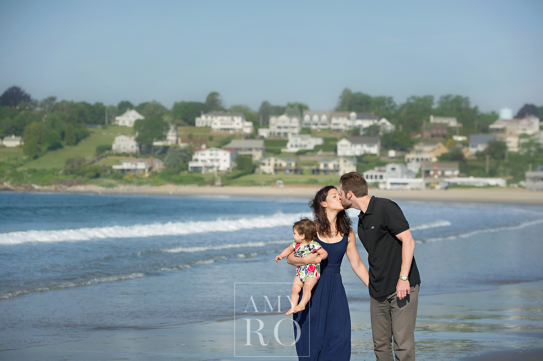 Color image of couple walking on beach with baby daughter kissing taken during family session at Bonnet Shores in Narragansett