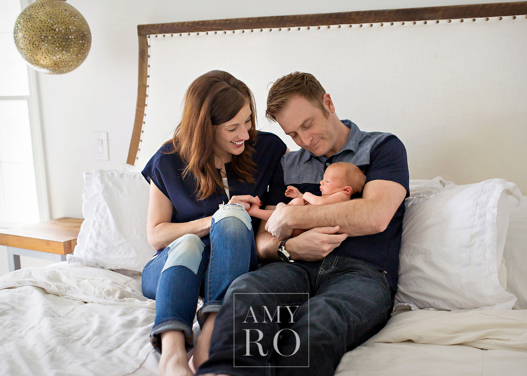 Image of mom and dad with newborn  snuggling on their restoration hardware bed  during an in-home lifestyle newborn session in East Greenwich