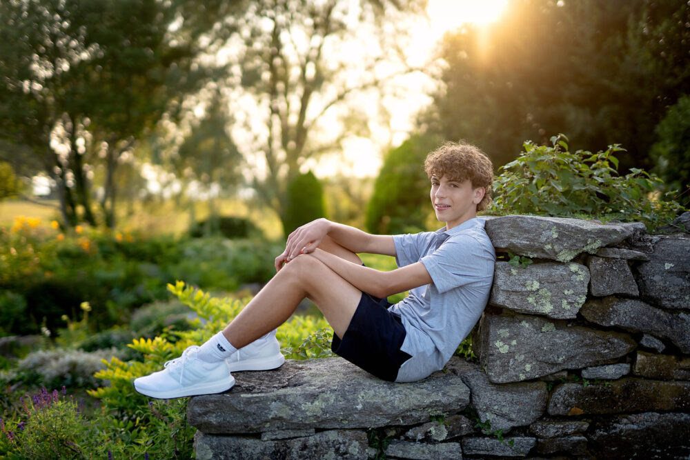 Backlit photo of Cheshire High School senior boy sitting on a stone wall in Connecticut